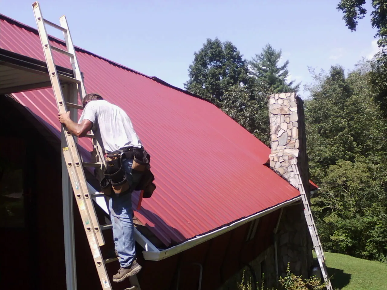 Roofer on a ladder beside a red metal roof and stone chimney — Soffit & Fascia Repair by Roofing Brothers in Torrington